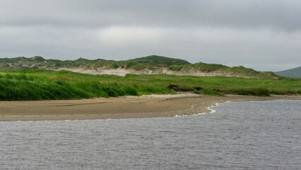 Serene landscape featuring a calm river flowing alongside lush green grass and sandy shores, with gentle hills rising in the background under a cloudy sky