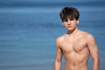 Shirtless confident teenager wearing earbuds enjoys music isolated on blue sea water in a sunny day...