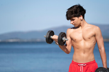 Shirtless athlete performs bicep curls with dumbbells, enjoying a workout by the tranquil sea