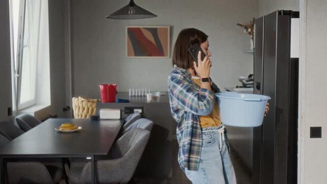 Medium shot of young woman calling plumber by phone while holding plastic bucket and looking up at leak in ceiling, copy space