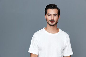 Confident young man with stylish beard wearing a white t-shirt stands against a solid grey background smiling at the camera. Portrait photography concept.