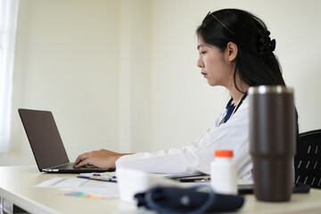 Young asian woman doctor in white uniform with stethoscope taking notes, using laptop, mobile...