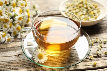Aromatic tea in glass cup and chamomile flowers on wooden table, closeup