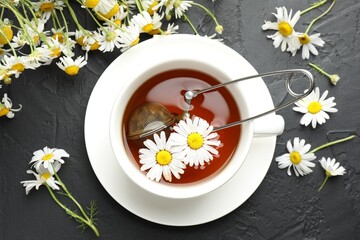 Tasty chamomile tea, infuser and flowers on black table, flat lay