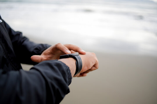 An athlete wears a smartwatch on his arm, is running and using an app. A male trainer in sportswear is on the beach.