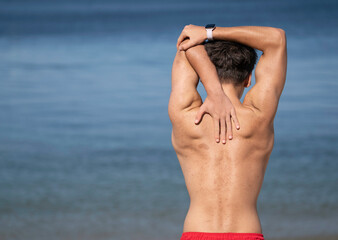 Shirtless swimmer stretches his back muscles on the beach before entering the ocean for a swim