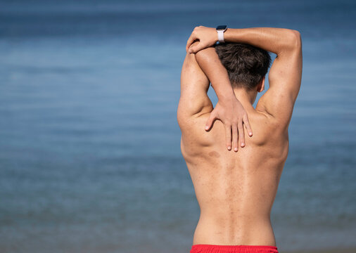 Shirtless swimmer stretches his back muscles on the beach before entering the ocean for a swim