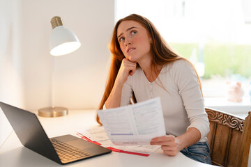 Woman reflects on a document while sitting at a desk with a laptop in a bright room, contemplating...
