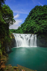 Fototapeta premium The breathtaking Shifen Waterfall plunges into an emerald pool, surrounded by lush green forests under a clear blue sky in Pingxi, New Taipei City, Taiwan, during summer.