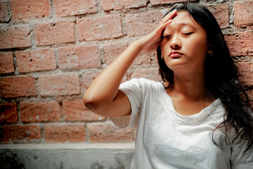 Young beautiful Indonesian teenage girl in casual white shirt leaning on brick wall, putting her hand on her forehead, having headache and feeling sick. Mental health and frustration concept.