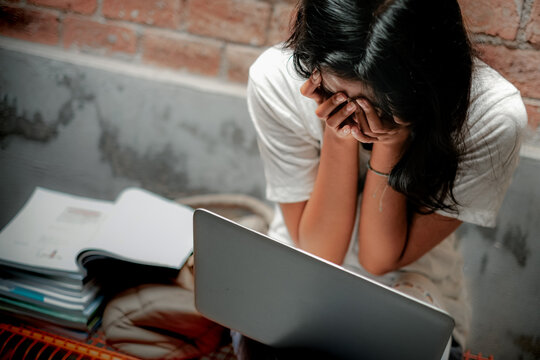 A young Asian teenage girl covering her face with both hands in front of a laptop while studying for high school final exam. Distress, frustration and mental health in education concept. - Powered by Adobe
