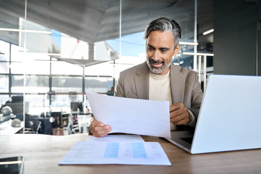 Middle aged professional business man company executive ceo bank manager lawyer wearing suit sitting at desk in office working checking corporate financial report documents using laptop.