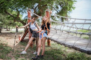 Two girls are standing on a bridge. One of them is wearing a backpack. They are both smiling. Two female friends hiking in nature, crossing a rope bridge on an adventure travel journey.