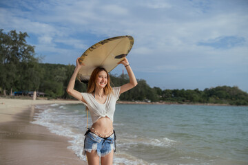 A woman is walking on the beach holding a surfboard. She is wearing a white shirt and blue shorts....