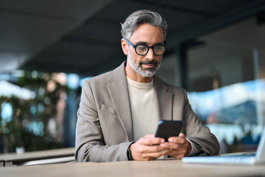 Stylish older busy businessman investor wearing glasses holding smartphone looking at cellphone doing financial payments. Happy middle aged business man using mobile phone sitting outside office. - Powered by Adobe