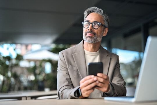 Happy business man executive of middle age using mobile phone sitting outside office. Stylish older busy businessman investor wearing glasses holding smartphone looking away with cellphone in hands. - Powered by Adobe