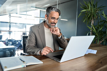 Naklejka na ściany i meble Busy mature older professional executive making video conference call. Smiling middle aged business man manager wearing earbud having virtual meeting looking at laptop hybrid working in office.