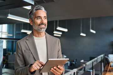 Happy confident mature business man executive standing in corporate office using digital tablet. Smiling middle aged professional businessman manager wearing suit looking away at work in modern space.