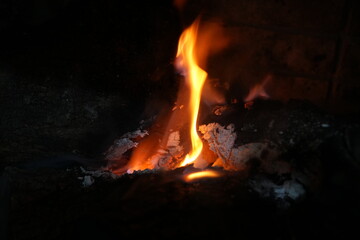 Close-up shot of a vivid orange flame rising from glowing embers and charred wood in a dark...