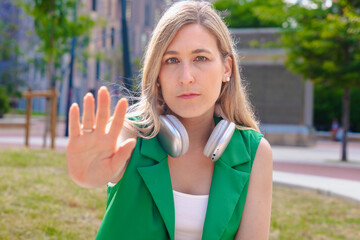 Serious young woman in green vest with silver headphones raises hand toward camera in stop gesture...