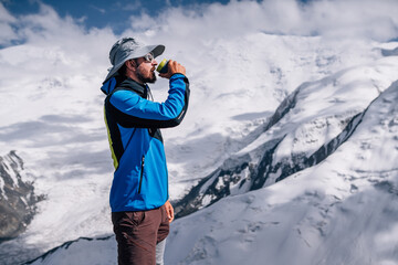 Climber in a hat drinking hot tea on Yuhin peak during Lenin peak ascent under mountain peaks in...