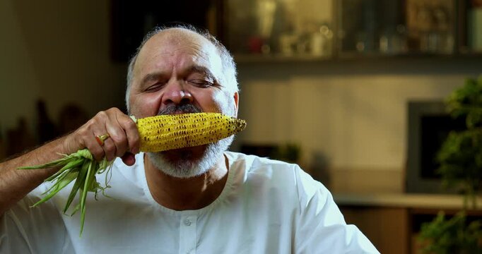 Senior Indian man eating roasted maize or bhutta at home in modern kitchen, Asian foodie old man expressing immense joy and happiness while enjoying healthy traditional Indian snack at dining table