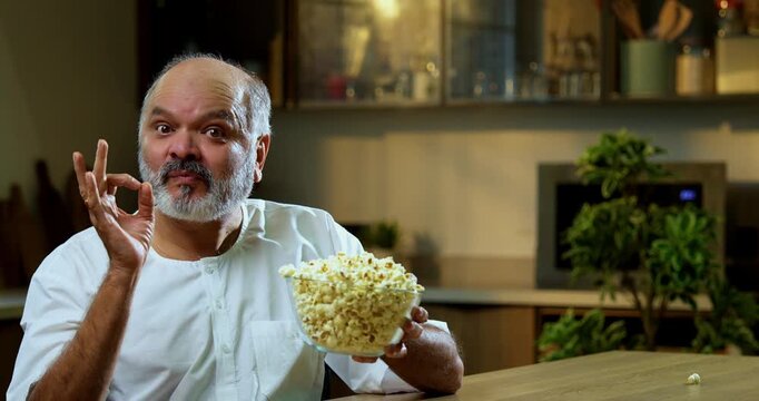 Senior Indian man eating popcorn from bowl in modern kitchen, Asian foodie old man expressing immense happiness while enjoying healthy snack, throwing popcorn in air, catching it by mouth playfully