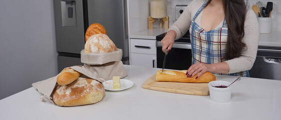 A person in a checkered apron slices a baguette on a wooden cutting board, with various bread bags and a plate of butter nearby. This highlights home baking and fresh food preparation
