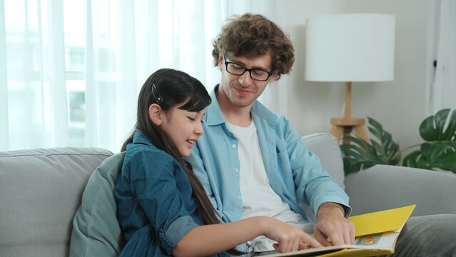 Happy daughter lean on father shoulder while listening story telling. Young asian schoolgirl looking ar dad while reading story book together and sitting at sofa. Family recreation concept. Pedagogy.