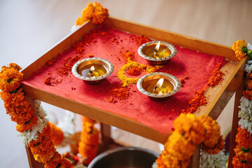 Three lit diyas and scattered red kumkum powder on a red cloth, decorated with marigold and jasmine garlands, signifying a hindu puja ritual