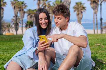 Young couple sitting on seaside lawn beneath palm trees, smiling as they browse a yellow smartphone...