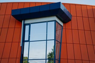 Modern orange facade with geometric blue-framed glass window reflecting sky and trees, contemporary...
