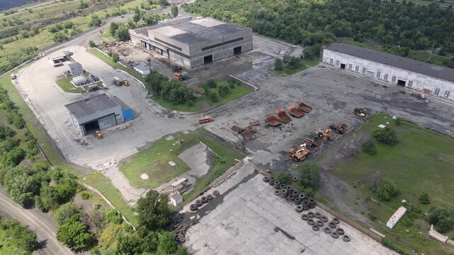 Aerial view of an abandoned industrial zone captured by drone. Old buildings, machinery, scattered tires, and overgrown greenery surround the deteriorating facility. High angle and wide coverage.