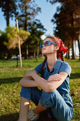 Naklejka premium young woman wearing blue sunglasses and red headscarf sitting on grass outdoors in sunny park with trees in background, relaxed and thoughtful mood