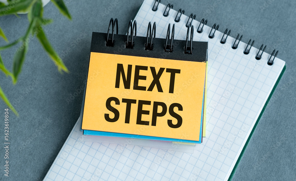 Poster a yellow sticky note with the word next steps text written on it. the note is on top of a spiral bou
