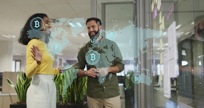 Two professionals in business attire analyzing holographic Bitcoin map on glass wall in open office