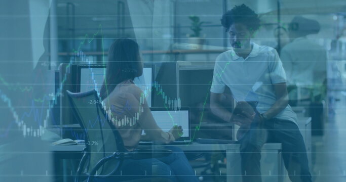 Colleagues analyzing monitor data and consulting tablet at office desk, overlaying financial charts