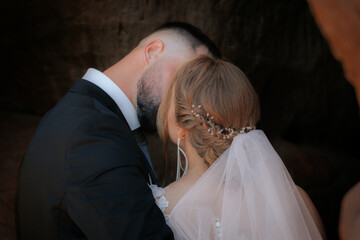 Joyful bride smiles as the groom kisses her cheek, framed by rocky cliffs. She holds a floral...