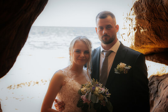 Bride and groom share a tender moment inside a sunlit cave by the beach, with ocean in the background and bouquet in hand. - Powered by Adobe