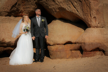 Wedding couple stands hand in hand near a carved sandstone rock wall, bathed in warm sunlight, with...