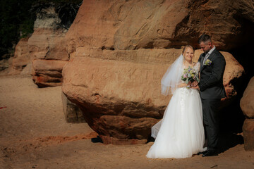 Wedding couple stands hand in hand near a carved sandstone rock wall, bathed in warm sunlight, with...