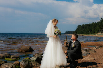 Groom kneels and gazes at the smiling bride standing on a rocky beach, surrounded by waves and a...