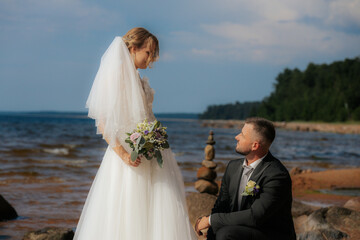 Groom kneels and gazes at the smiling bride standing on a rocky beach, surrounded by waves and a...
