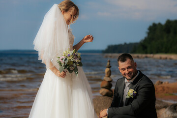 Groom kneels and gazes at the smiling bride standing on a rocky beach, surrounded by waves and a...