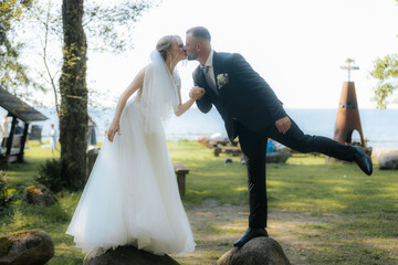 Bride and groom kiss while balancing on rocks in a playful pose near a lakeside, with trees and a scenic background adding romantic charm.