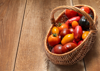 A wicker basket filled with colorful fresh tomatoes on a wooden background. A variety of types of tomatoes, including red, yellow, orange