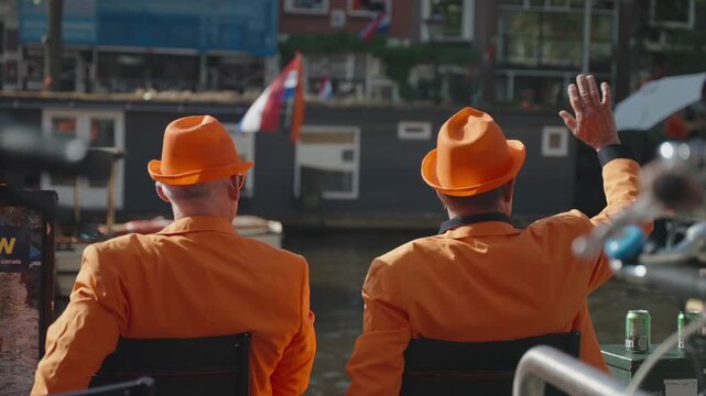 King's Day in Amsterdam. Two men near Channel in Orange Suites are Waving to Passing by Boats on Sunny Dutch Holiday.