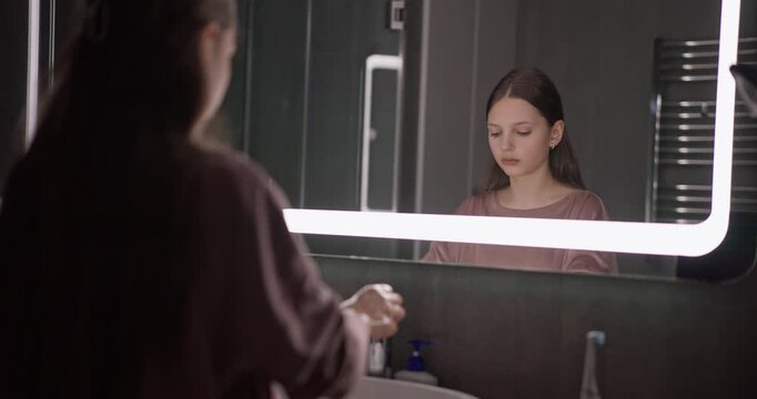 Teenage girl washing her hands and drying them with towel while standing in front of backlit mirror in modern bathroom at home
