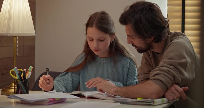 Teenage girl doing homework with her dad support sitting at table with desk lamp at home in the evening. Young father and daughter doing homework in the kitchen at home