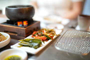 Plates of kalbi, soup, kimchi, namul and grilled meat glow on table in a busy barbecue restaurant,...